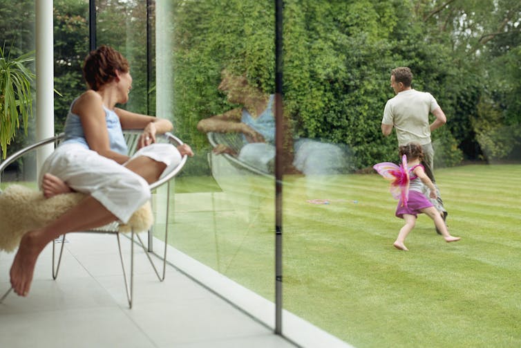 woman looks out of home's large windows at girl playing with man in the garden