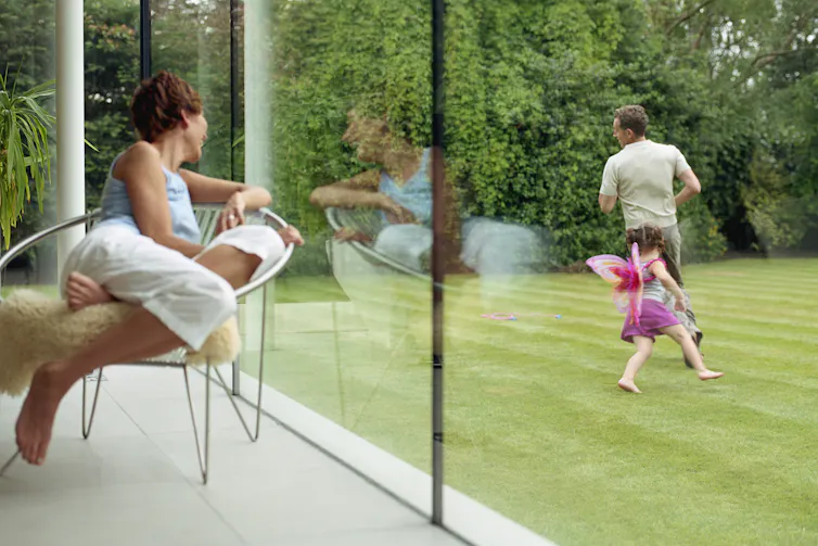woman looks out of home's large windows at girl playing with man in the garden