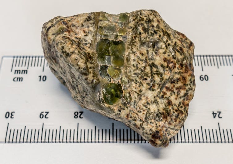 A small, irregular tan rock with translucent green crystals in it sitting on a metric ruler.