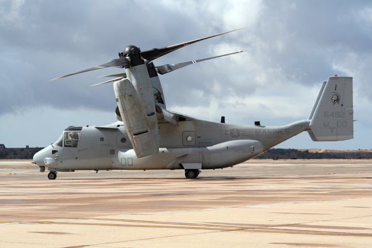 A sand coloured tarmac and a white plane with two rotors on top seen from the side