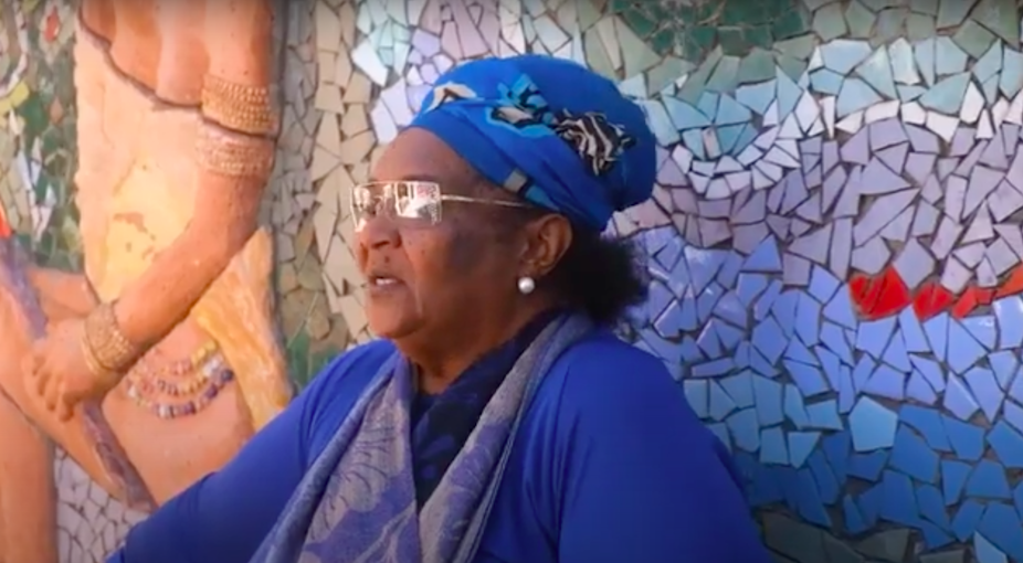 A woman in a headscarf and a blue outfit is pictured in profile against a mosaic mural backdrop.