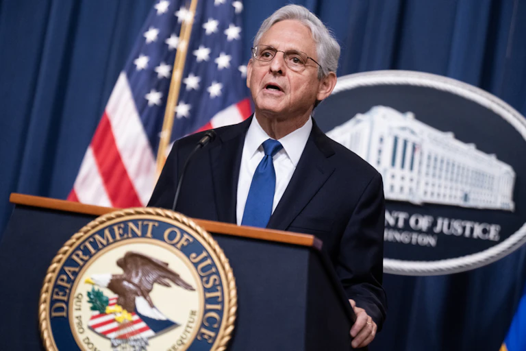 A man in a coat and tie stands at a lectern in front of a U.S. flag.