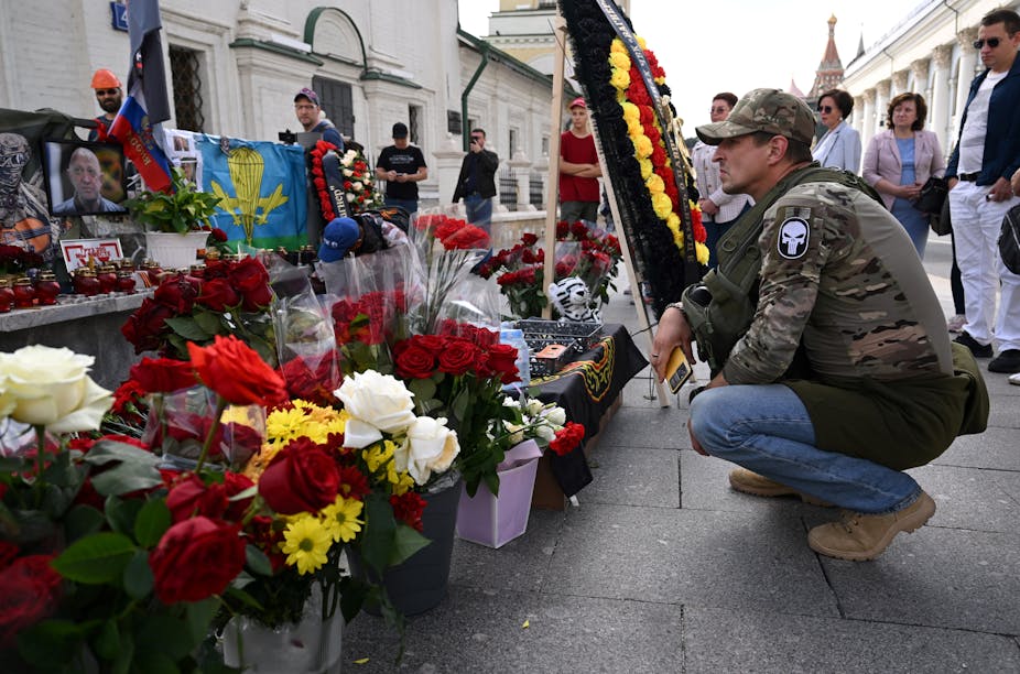 Homme accroupi devant un mémorial de fleurs et de photos parmi lesquelles celles d'Evguéni Prigpjine et de Dmitri Outkine