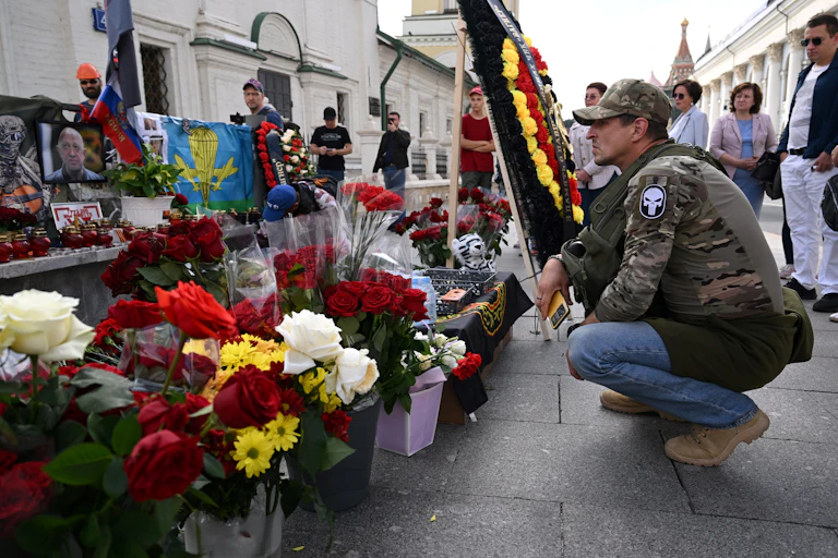Homme accroupi devant un mémorial de fleurs et de photos parmi lesquelles celles d'Evguéni Prigpjine et de Dmitri Outkine
