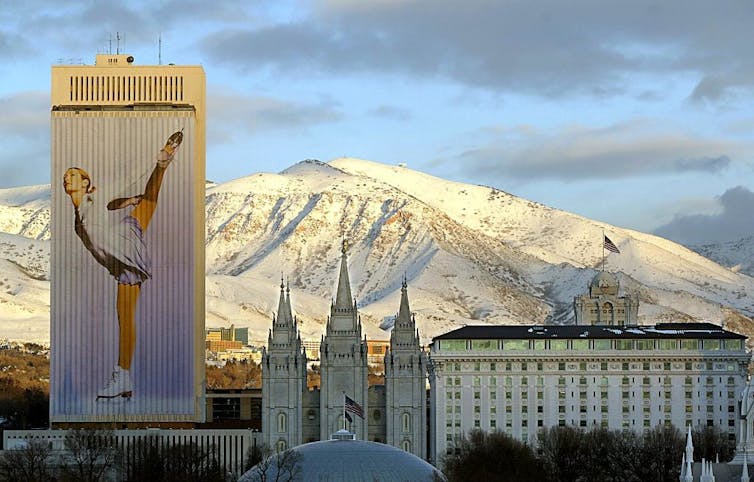 A tall building with an image of a woman figure skating projected on it, next to church steeples and a snow-covered mountain.