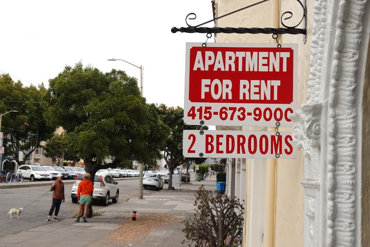 A 'for rent' sign for a 2 bedroom apartment is hung outside of a building on a quiet street.
