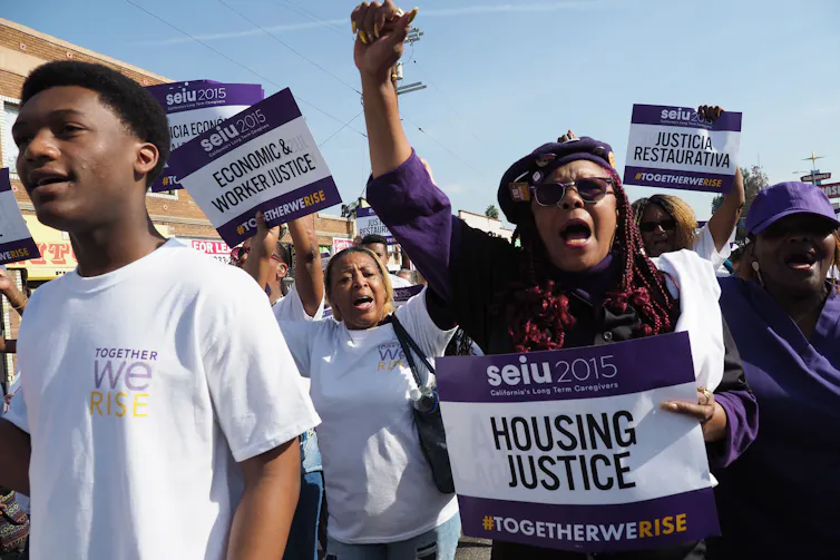 A crowd of Black people hold signs with purple that say 'SEIU 2015' and 'Economic and worker justice' and raise their hands in the air.