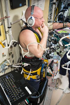 A bald man in a white headset and tight, dark pants with many wires attaching them to surrounding machinery.