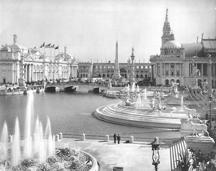 A black and white photo of ornate buildings around a waterway, with fountains in a plaza.