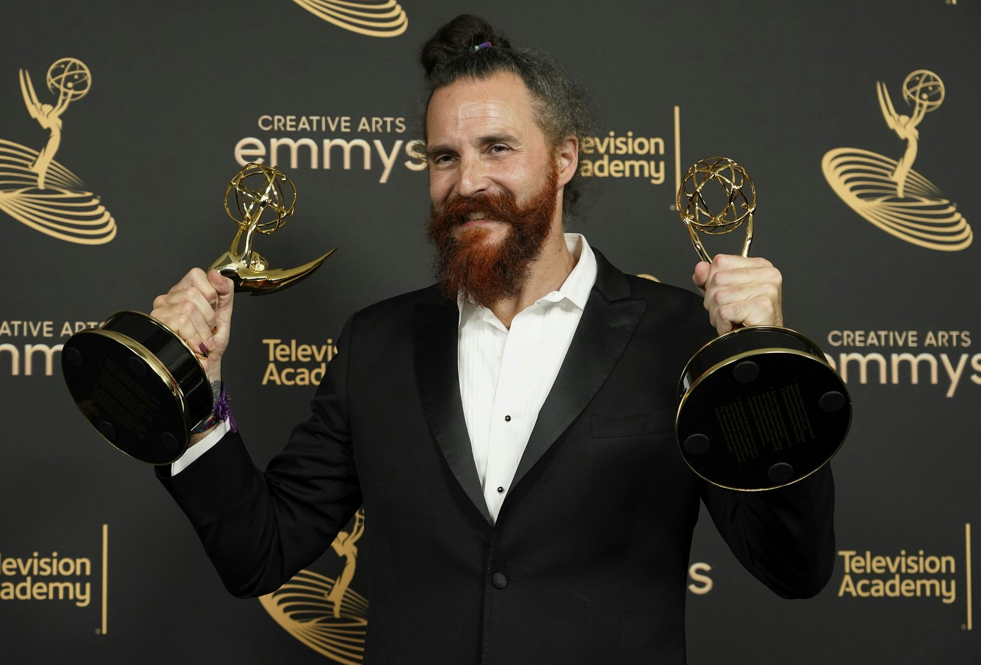 A man in white shirt and black jacket holding two awards.
