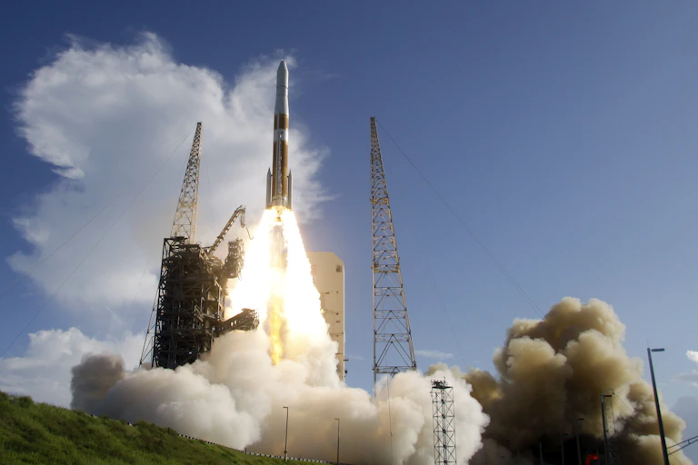 A white and gray rocket launches with a cloud of smoke, on a partly cloudy day.
