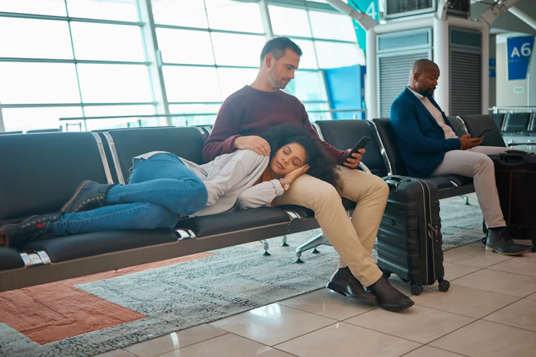 Woman lying on a row of chairs beside two men with suitcases looking at phones, row of windows in the background.