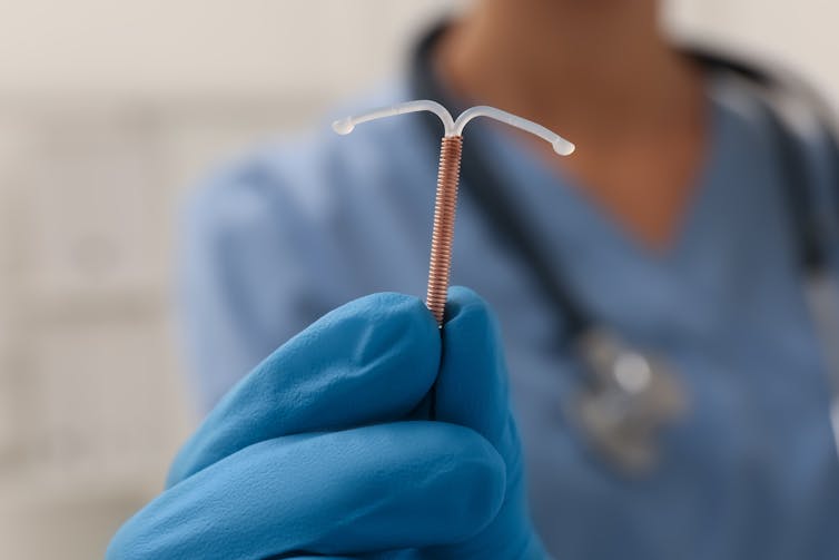 A clinician wearing blue surgical gloves holds the copper IUD in their hands.