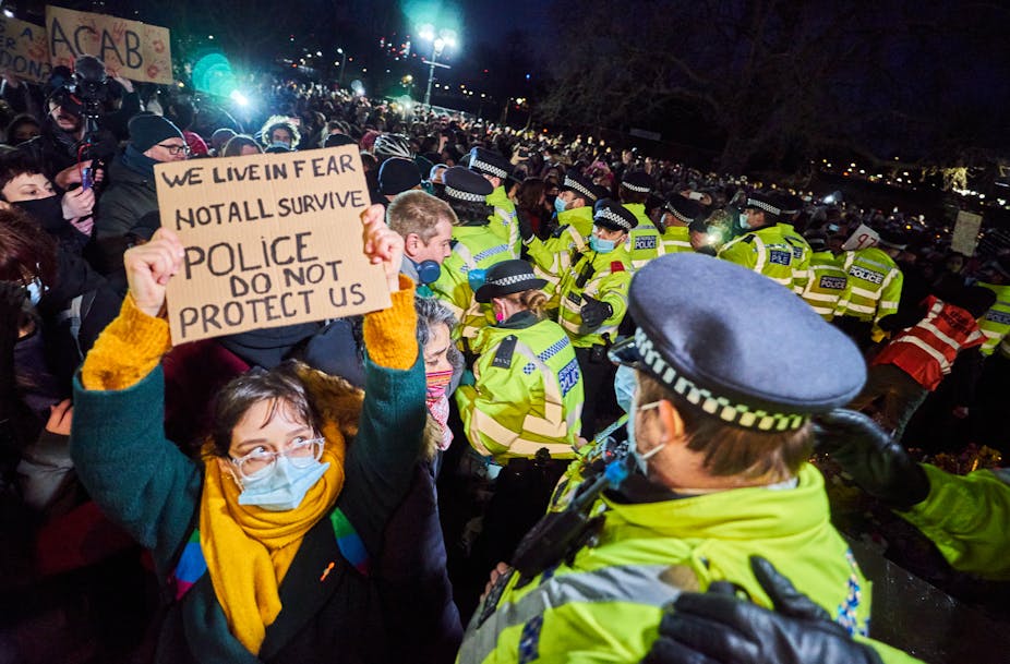 Amid a crowd of Met police officers during a nighttime protest, a woman holds a cardboard sign reading 'We live in fear, not all survive, police do not protect us'