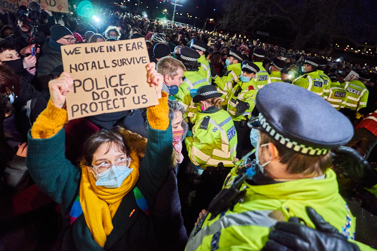Amid a crowd of Met police officers during a nighttime protest, a woman holds a cardboard sign reading 'We live in fear, not all survive, police do not protect us'