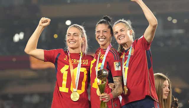 Tres mujeres vestidas con la camiseta de España de fútbol y portando medallas sonríen.