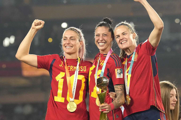 Tres mujeres vestidas con la camiseta de España de fútbol y portando medallas sonríen.