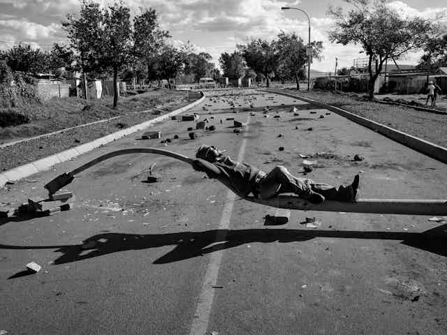 A man lies stretched out on the curve of a streetlamp that has been flattened, resting on a road. Behind him a street filled with concrete and signs of violence.