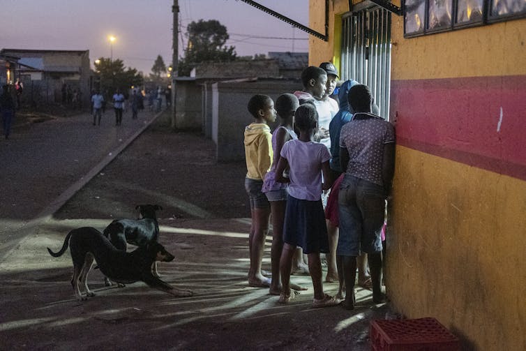 Children crowd the metal bars of a shop, light shining from inside it.