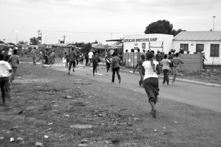 People run, their backs to the camera, fleeing during a protest in a township setting.