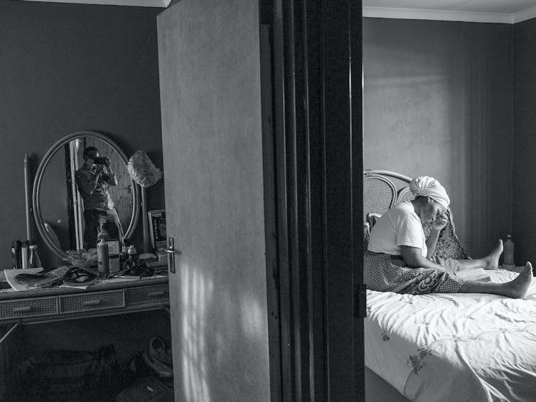 On one half of an image a woman sits reading on a bed, her legs splayed. In the other half a young man is elected in a mirror, taking a photograph of her.