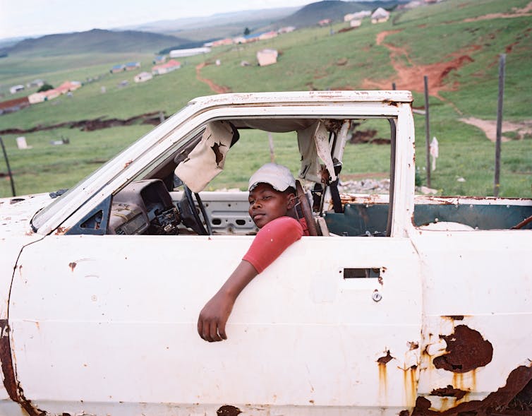 A rural setting with a young man seated in a rusty white car, his arm hanging out the open window and looking wryly at the camera.