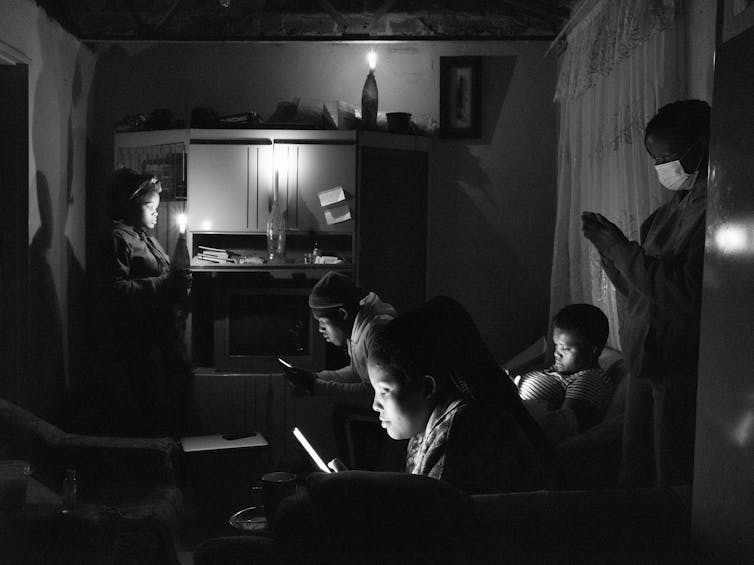 A darkened room with candles in cold drink bottles. Several people sit and stand, all staring into their phone screens, their faces lit by the screens.