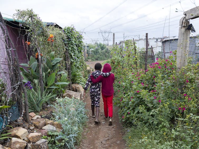 From behind, two young girls walk down a path, arms around one another. On either side of them are greens and blooming flowers.