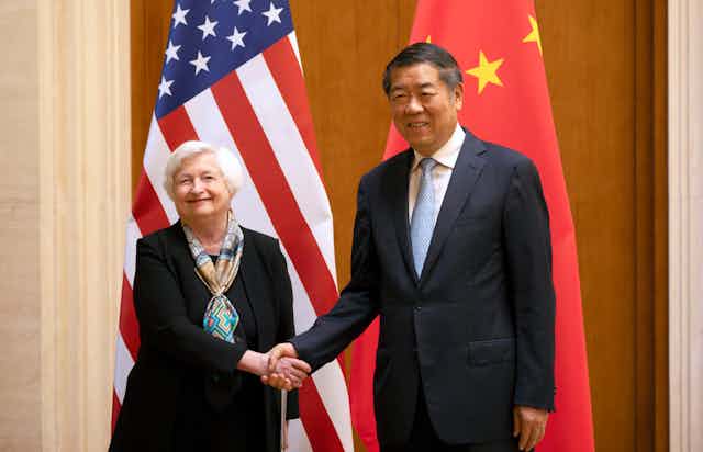 A white-haired woman shakes hands with a taller man. American and Chinese flags are behind them.