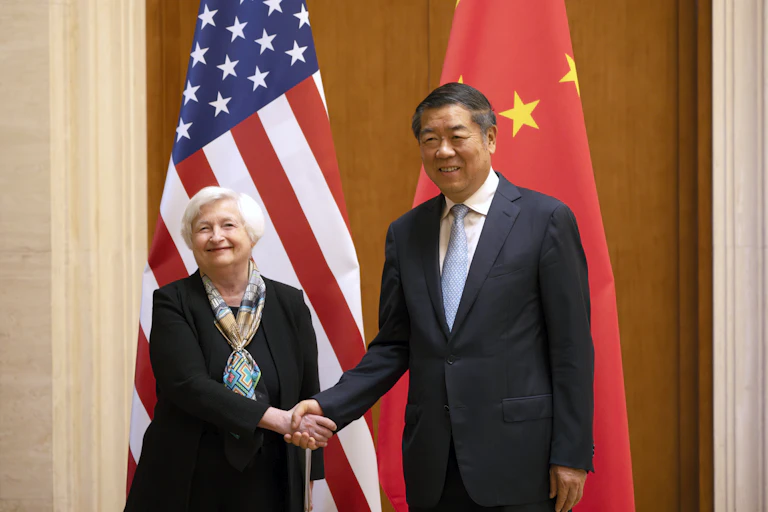 A white-haired woman shakes hands with a taller man. American and Chinese flags are behind them.