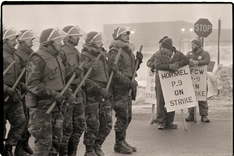 A large group of National Guardsmen in battle attire pass a small group of workers with signs indicating that they are striking Hormel employees in an old black and white photo.