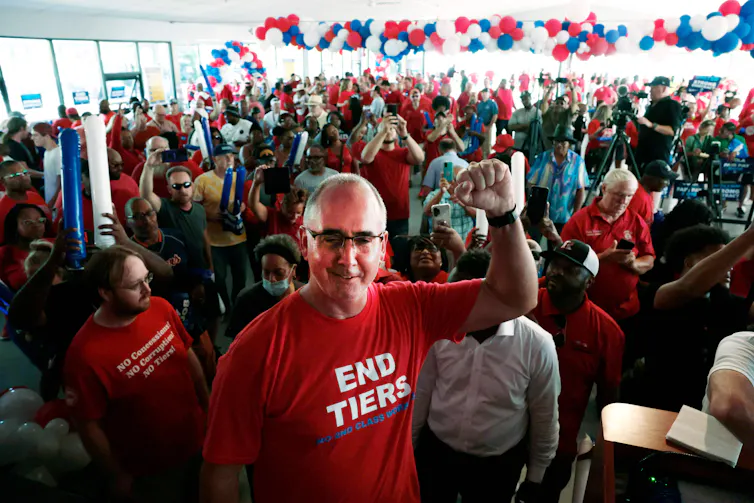 Man in a red t-shirt that says 'no tiers' holds his fist in a labor salute in fronb of a crowd of people dressed in red, white and blue.