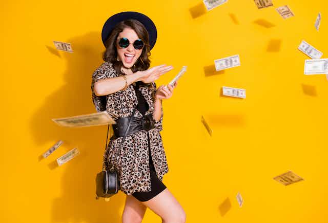 A young woman in sunglasses and a hat smiles and tosses money out of her hands, she is against a bright yellow background and surrounded by falling notes