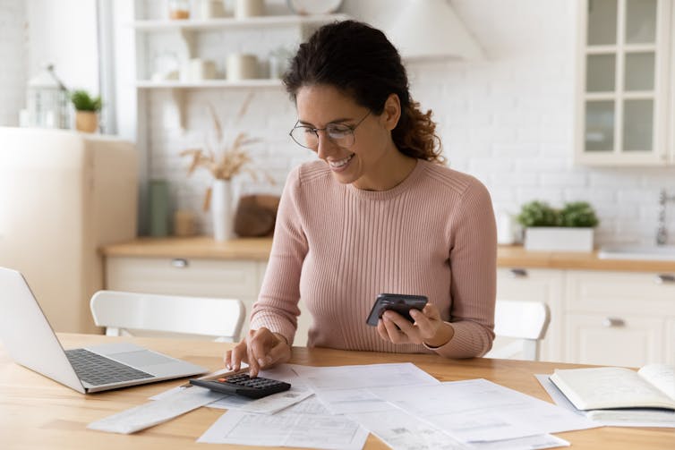 A young woman sits at a kitchen table with an assortment of financial papers in front of her, she smiles while typing into a calculator and computer