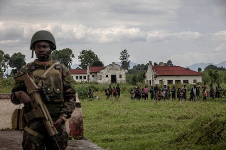 Un soldat tient un fusil tandis qu'un groupe d'enfants s'aligne derrière lui devant des bâtiments blanchis à la chaux qui se dressent sur un grand terrain herbeux.