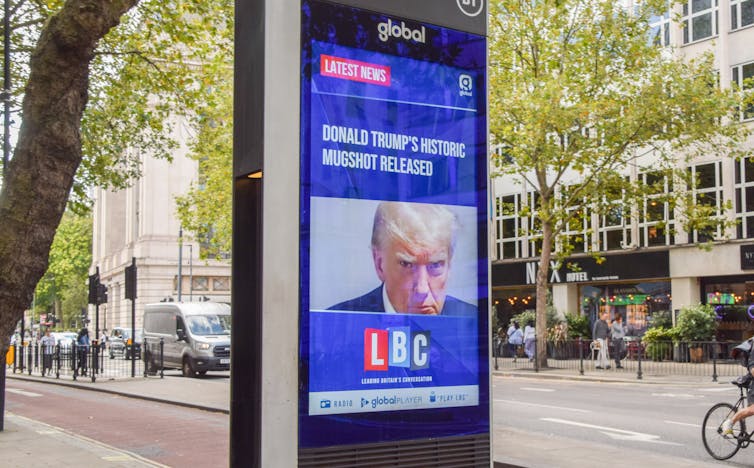 A large screen on a street with trees and parked cars shows a photo of Donald Trump's mugshot.
