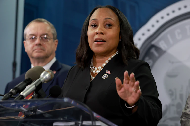 Fani WIillis wears a dark blazer and raises her hand at a lectern, standing next to a white man with glasses and a suit.