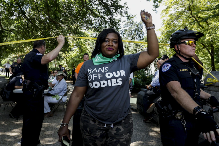 With police behind her, a woman wearing a T-shirt that reads “Bans off our bodies,” holds her left fist in the air.