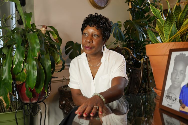 A Black woman, surrounded by large plants, sits with both hands resting on her crossed legs as she stares ahead.