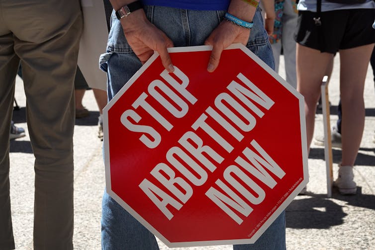 A person holds a red and white site, shaped like a stop sign, that reads: