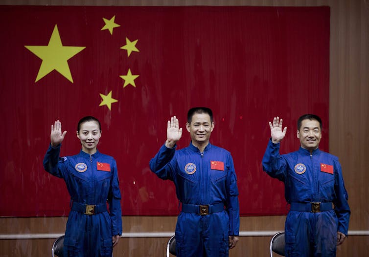 An Asian woman and two Asian men in blue jumpsuits smile and wave standing in front of a large red and gold chinese flag.