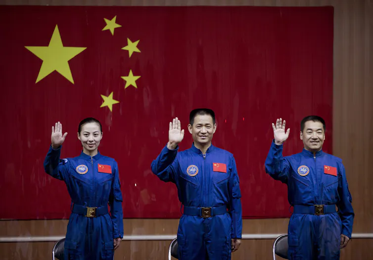 An Asian woman and two Asian men in blue jumpsuits smile and wave standing in front of a large red and gold chinese flag.