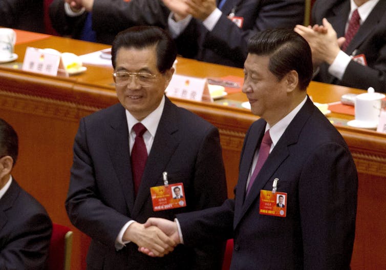 Two men in dark suits with red ties shake hands.