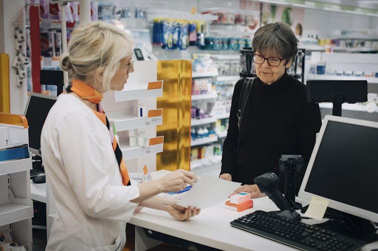 Older adult customer standing at a pharmacy checkout stand, with pharmacist explaining something.