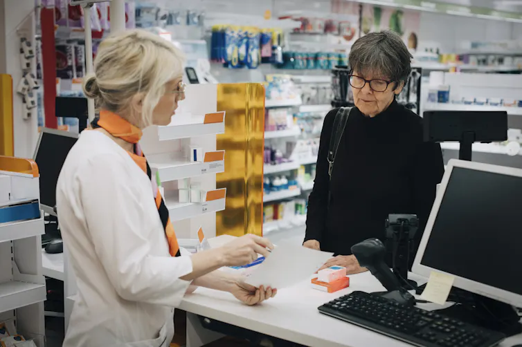 Older adult customer standing at a pharmacy checkout stand, with pharmacist explaining something.