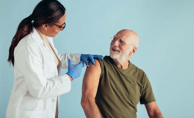 An older man in a green t-shirt getting a injection from a female doctor in a white coat and blue gloves