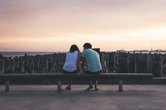 pria dan wanita muda duduk di tepi pantai sambil mengobrol saat matahari terbenam