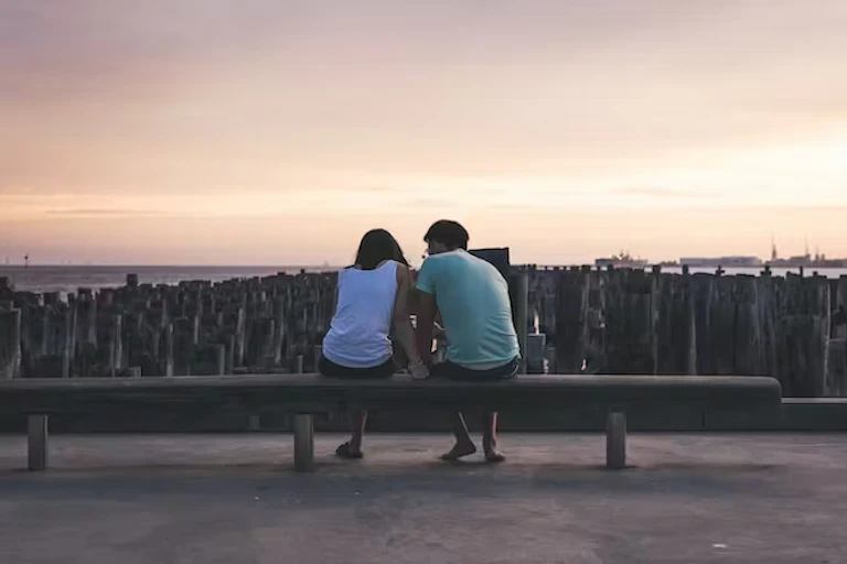 pria dan wanita muda duduk di tepi pantai sambil mengobrol saat matahari terbenam