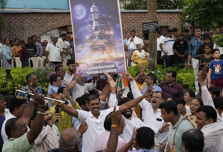 A group of cheering, smiling people hold signs depicting the Chandrayaan-3 lander.