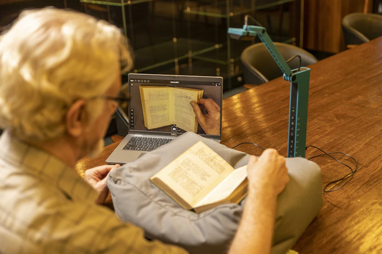 Homem folheia um livro na frente de um computador